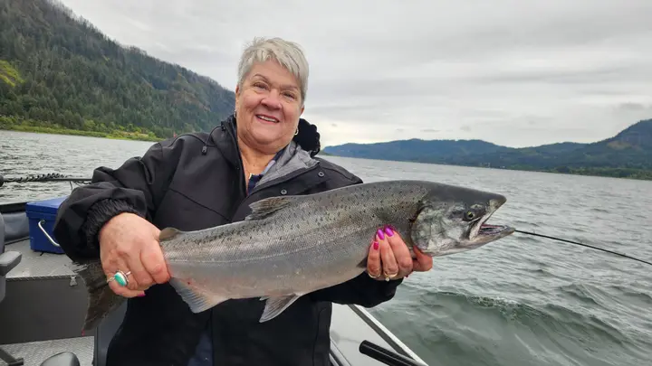 An older woman holding a coho salmon with the Columbia Gorge behind her