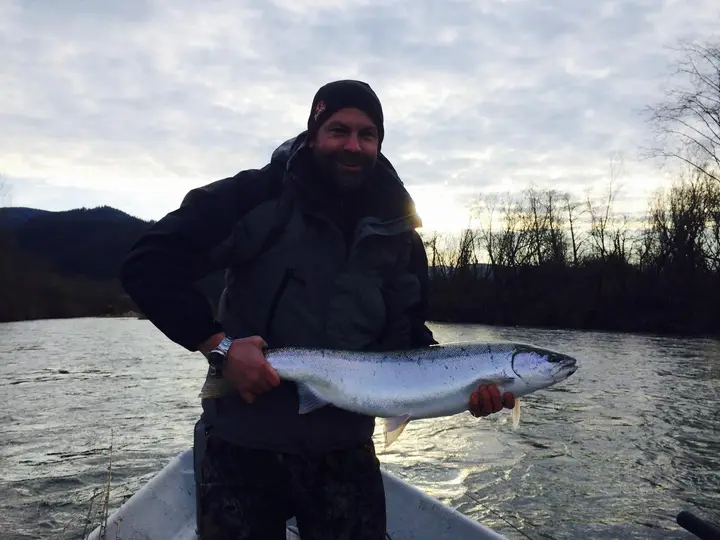 A man holding a winter steelhead beside a drift boat on a moody river