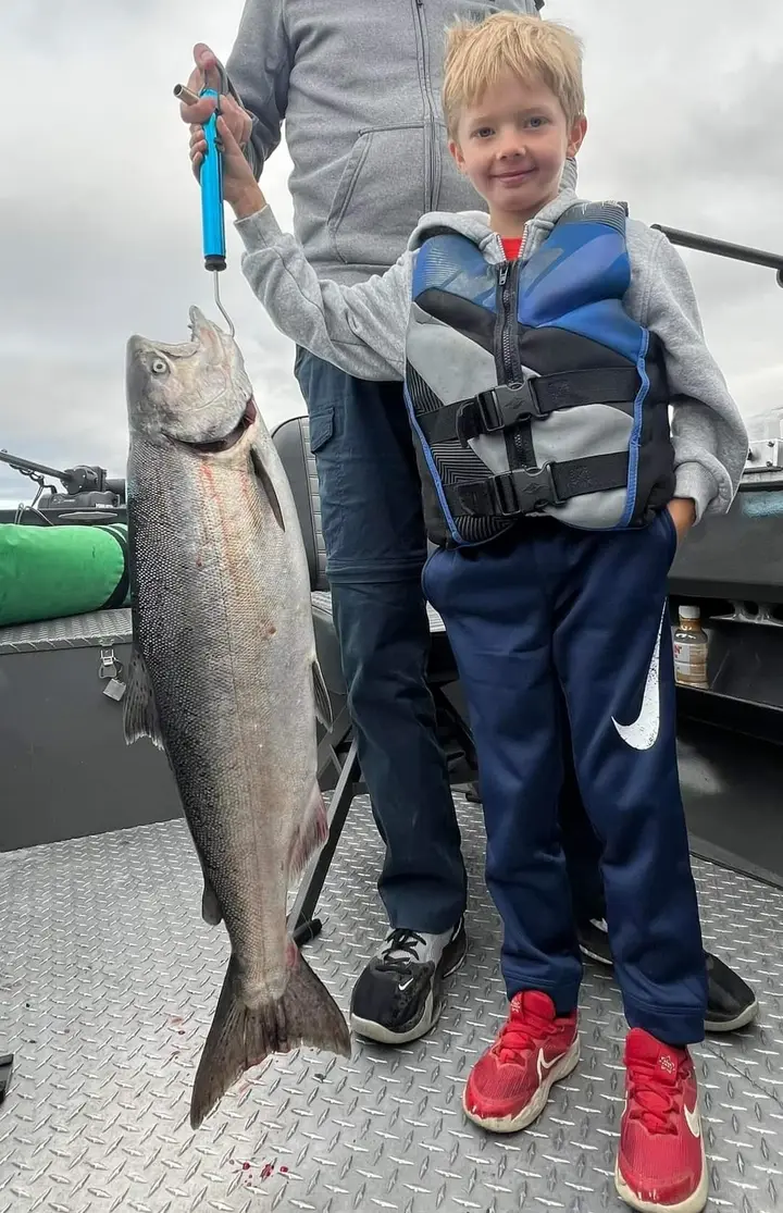 A small boy in a life jacket standing beside a hanging chinook salmon