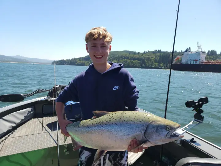 A teenage boy with a chinook salmon and a cargo ship passing behind